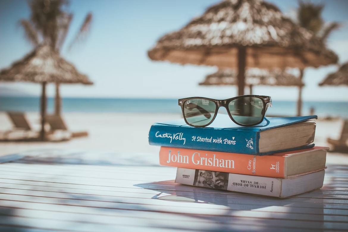 Paire de lunettes de soleil Ray Ban noires posée sur une pile de livres devant la plage avec des parasols et des transats