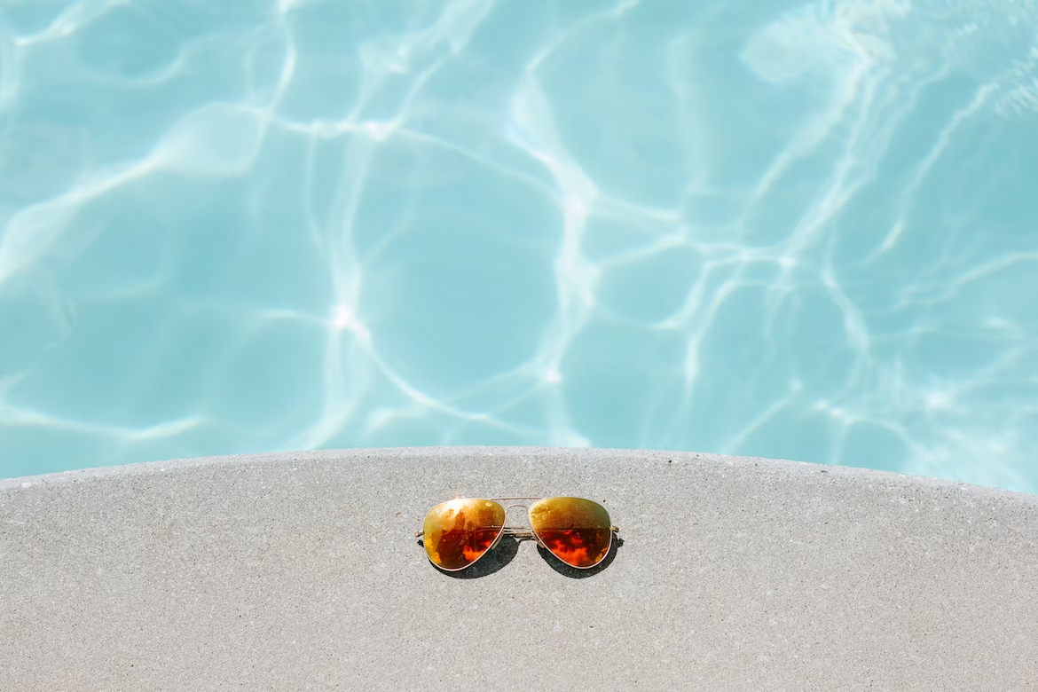 Paire de lunettes de soleil aviateur avec des verres irisés sur une dalle blanche devant une piscine