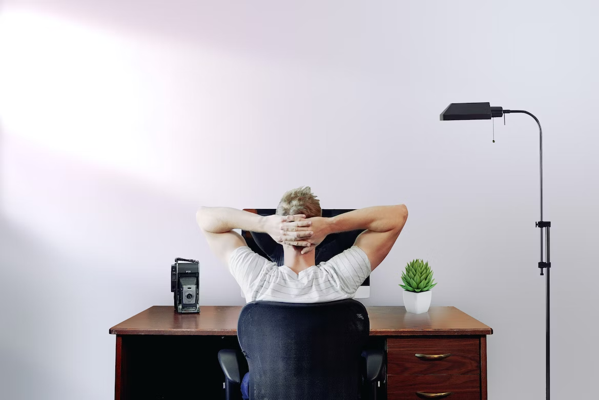 Homme de dos en T-shirt blanc rayé croisant les bras derrière la tête assis à son bureau devant son ordinateur.