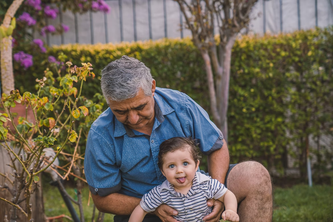 Un homme âgé en chemise bleue à manches courtes tient un bébé en polo rayé dans un jardin 