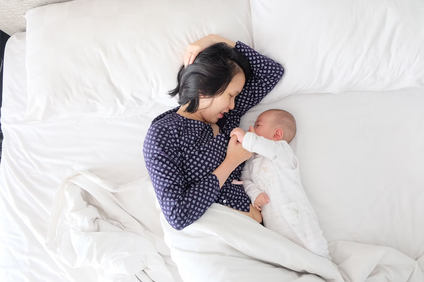 Femme en chemise bleue à motifs blancs couchée dans un lit deux places tenant la main d'un bébé