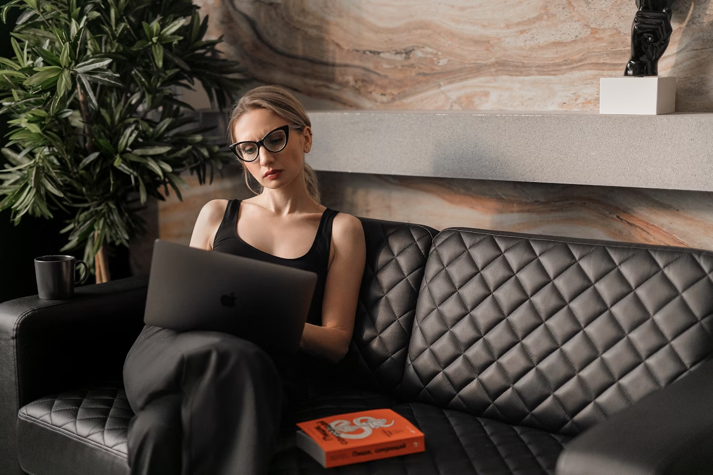 Femme en débardeur noir et avec des lunettes de vue travaillant sur un ordinateur assise sur un canapé en cuir noir