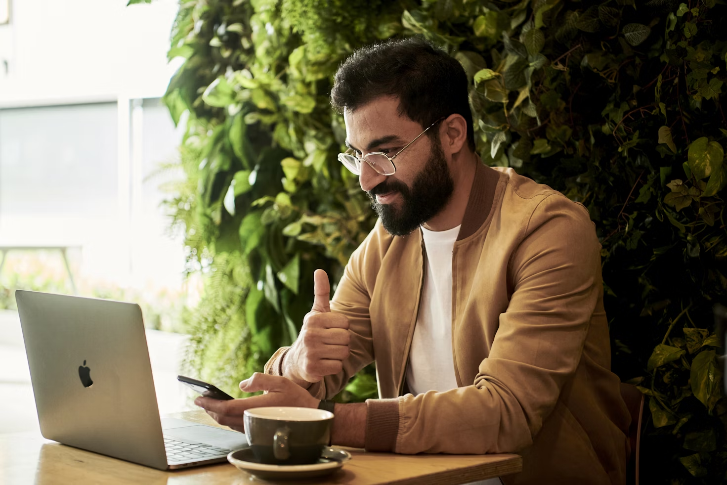 Homme avec une veste marron levant le pouce devant l'écran de son ordinateur portable posé à côté d'une tasse à café