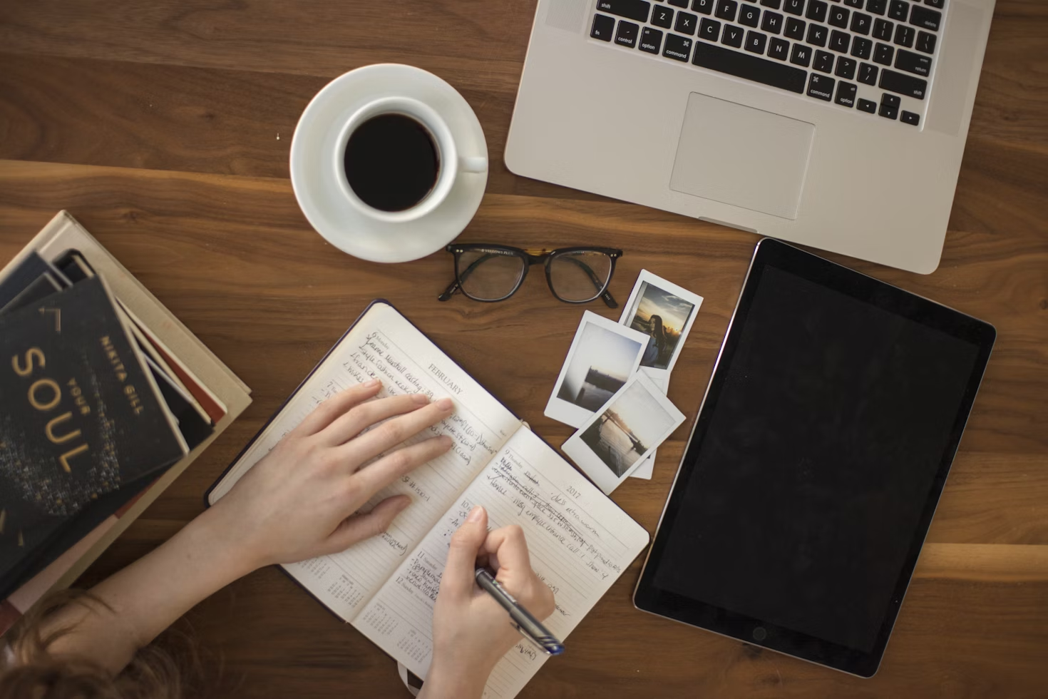 Personne écrivant dans un carnet sur une table en bois avec des polaroïds posés à côté d'une tablette de lunettes d'une tasse à café et d'un ordinateur