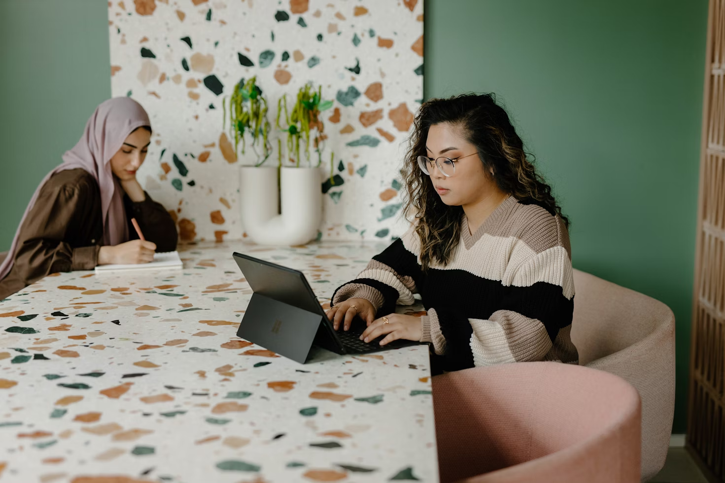 Deux femmes travaillant sur un ordinateur et écrivant dans un carnet assises à une table de réunion en pierres blanches vertes et roses