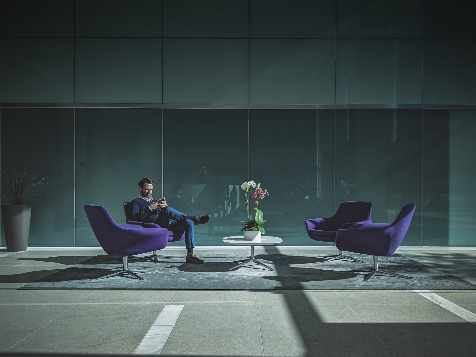 Homme assis sur un fauteuil violet devant une table basse avec un bouquet de fleur et regardant son téléphone