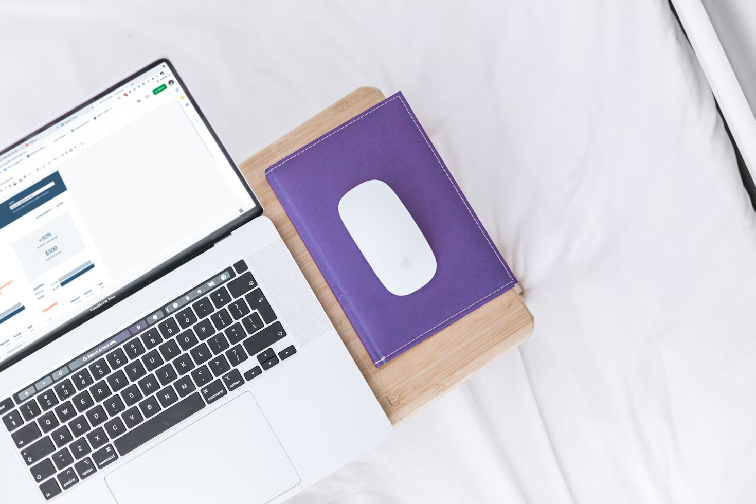 Ordinateur portable posé sur un lit à côté d'une souris bluetooth posée sur un carnet violet sur une tablette en bois.