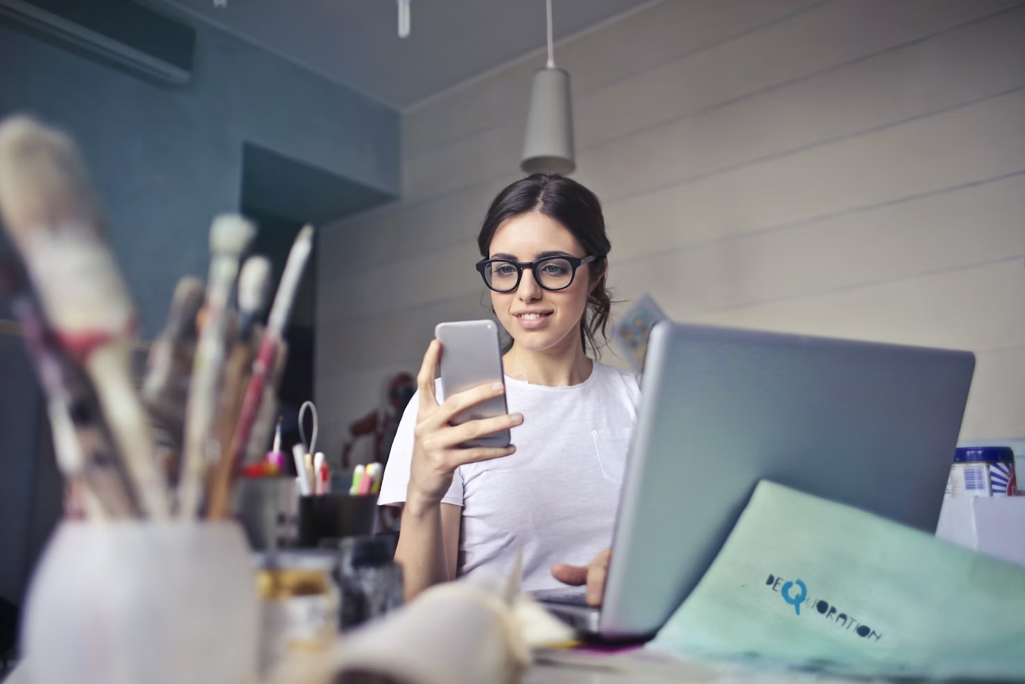 Femme en T-Shirt blanc et portant des lunettes assise devant un ordinateur portable et regardant son téléphone