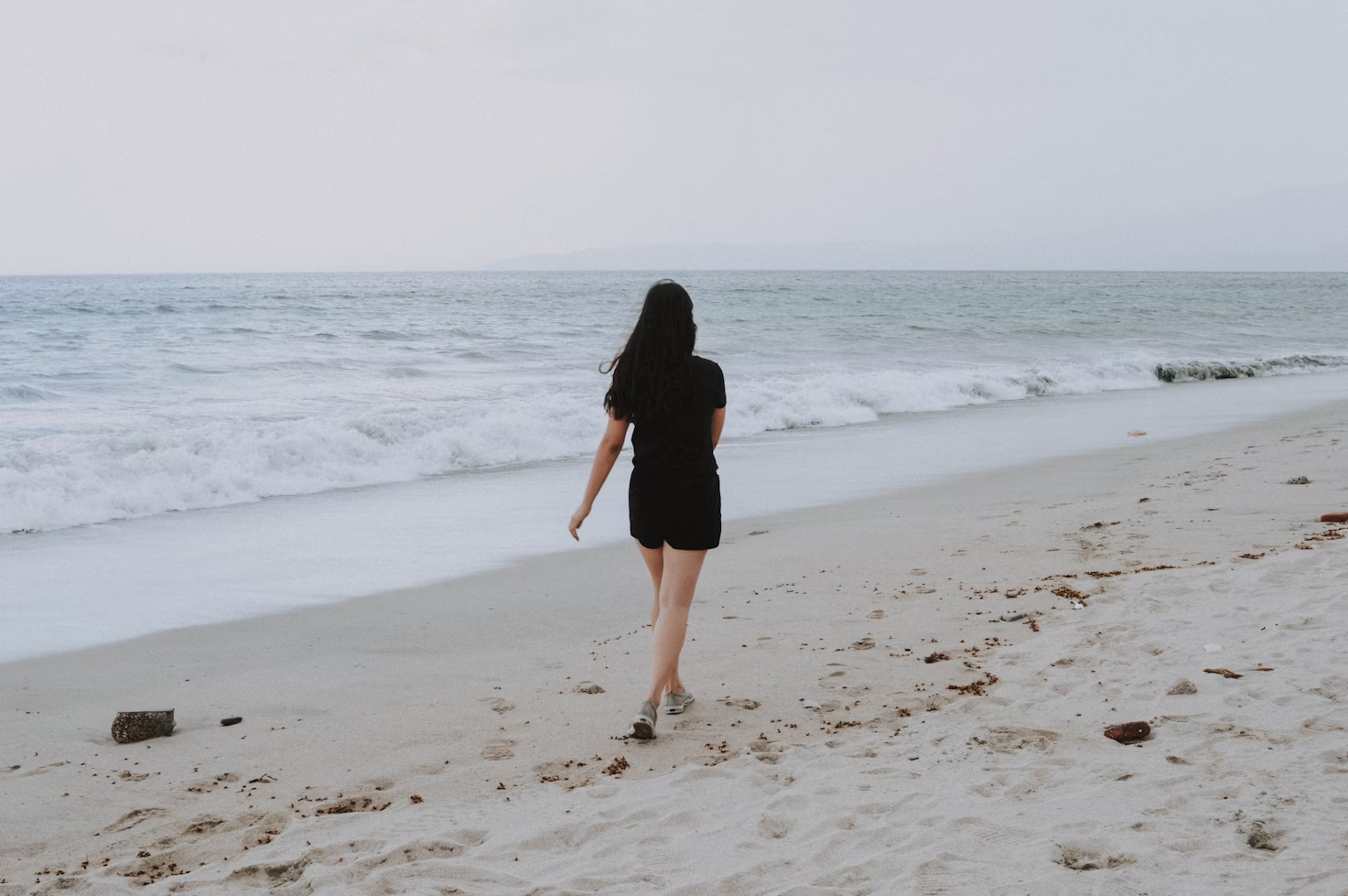 Femme avec des cheveux bruns un haut et un short noirs marchant sur une plage de sable blanc au bord de la mer