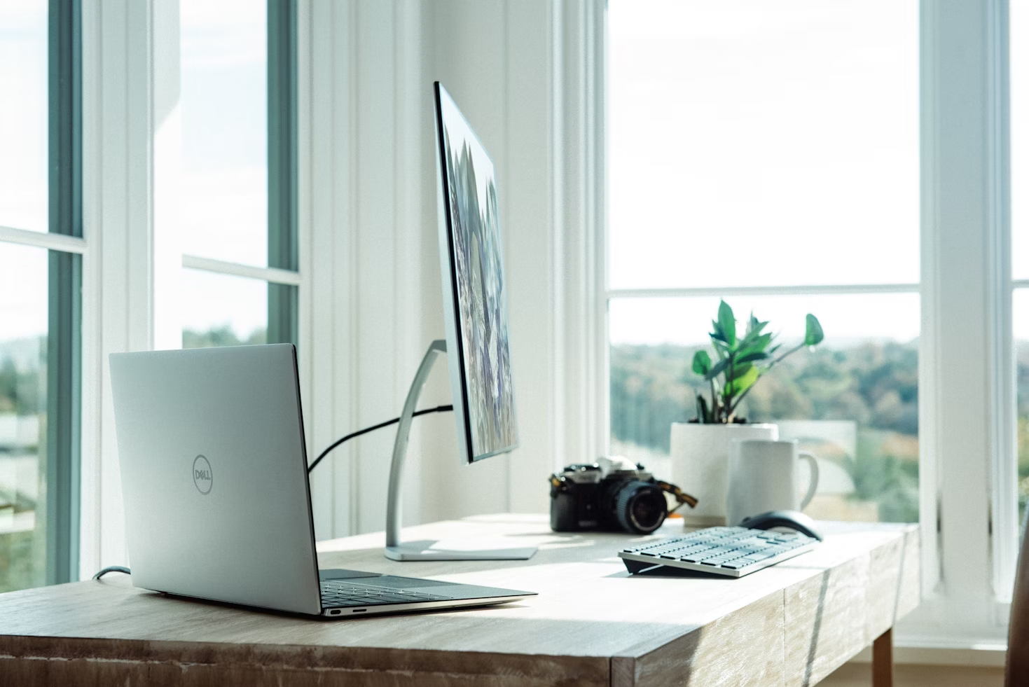 Bureau en bois clair avec un ordinateur portable un écran d'ordinateur et un clavier bluetooth devant une fenêtre