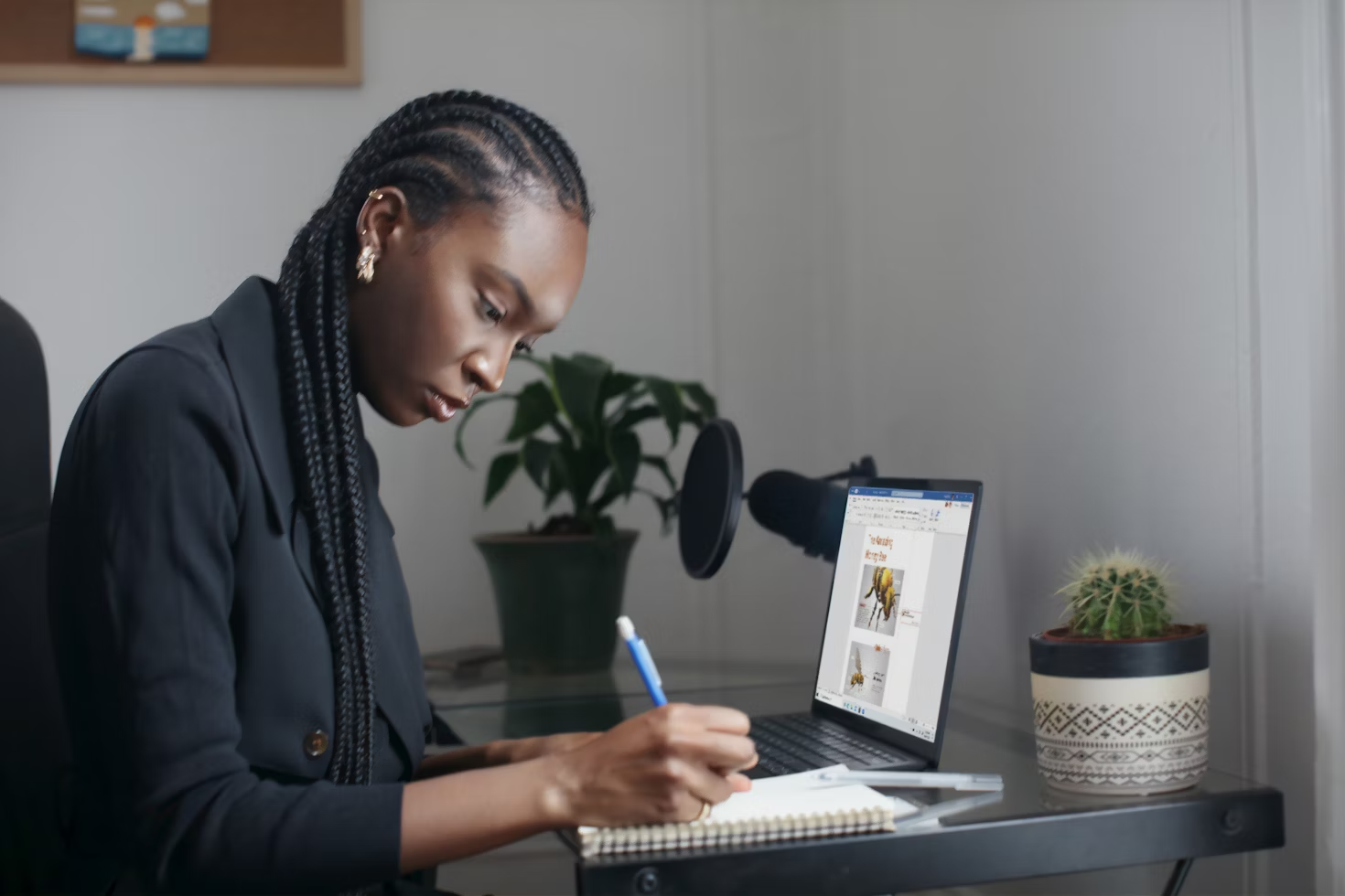 Femme écrivant au criterium sur un carnet à spirale assise à un bureau en verre devant un ordinateur à côté de plantes