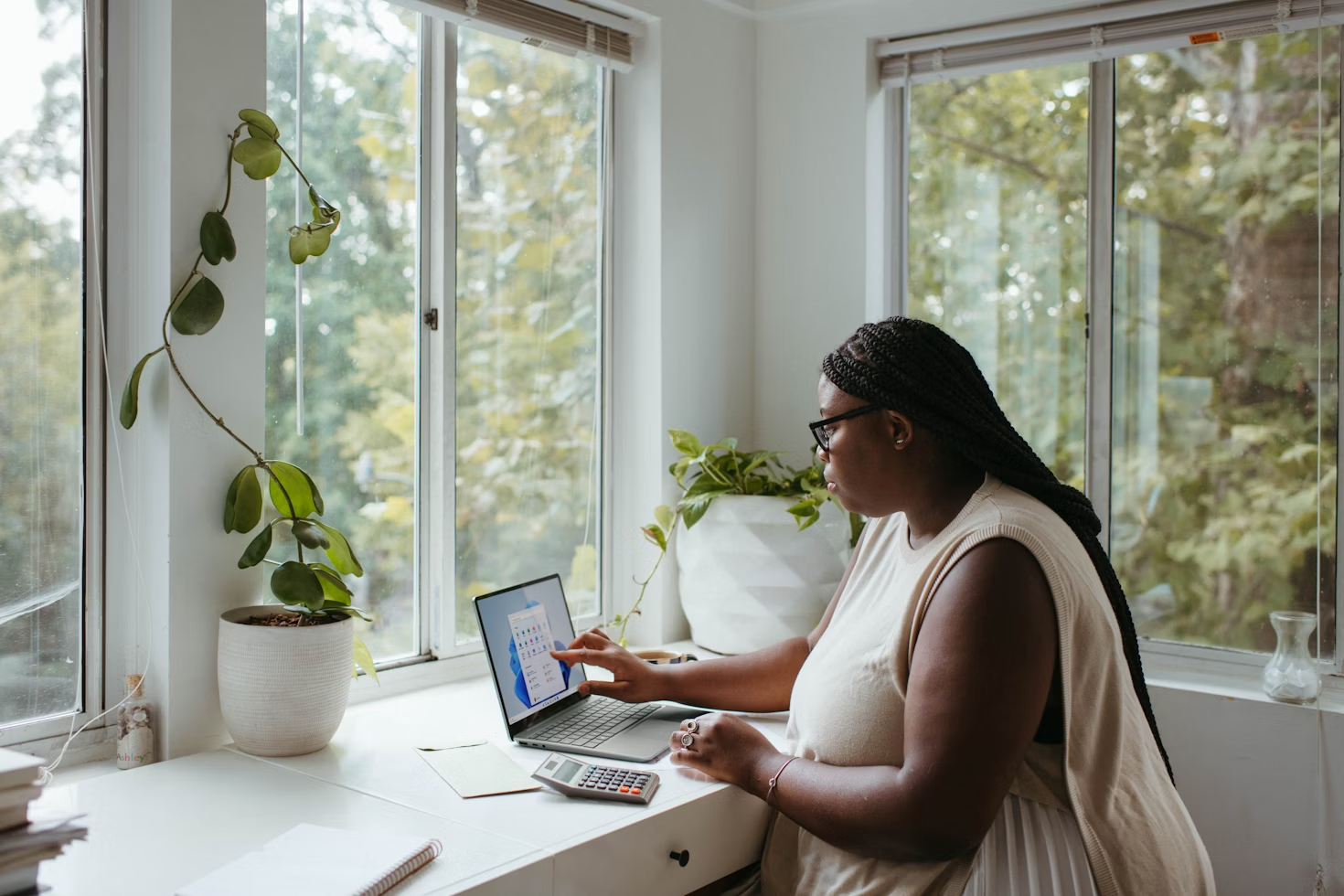 Femme avec des lunettes de vue noires travaillant sur son ordinateur portable posé sur un bureau blanc face à une fenêtre