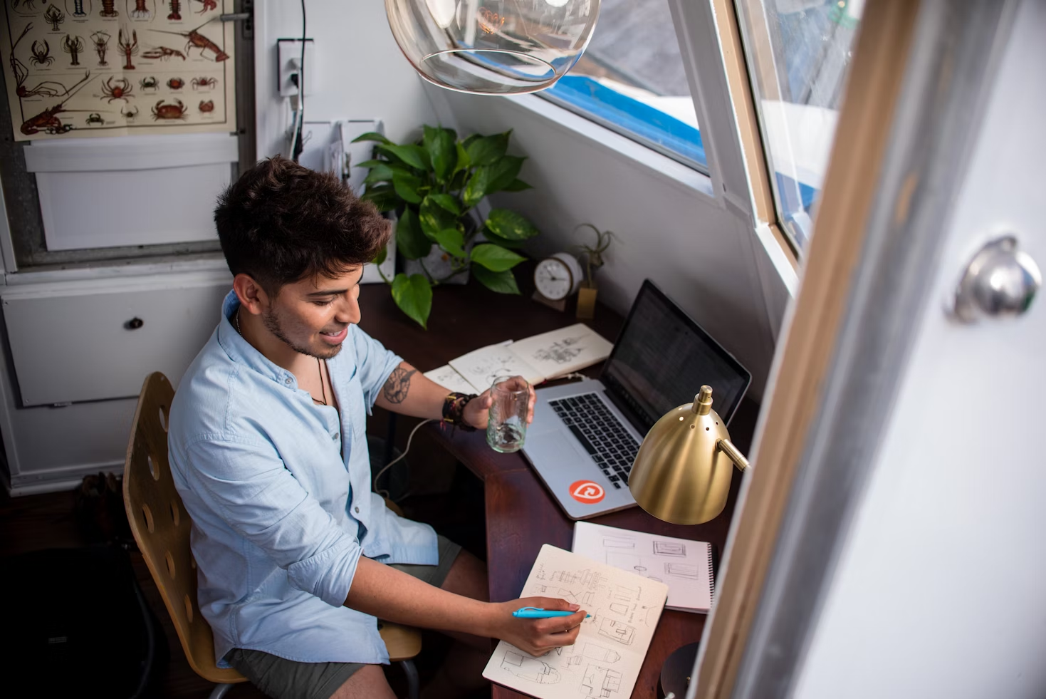 Homme portant une chemise bleue assis à un bureau devant un ordinateur portable tenant un verre dans sa main et écrivant