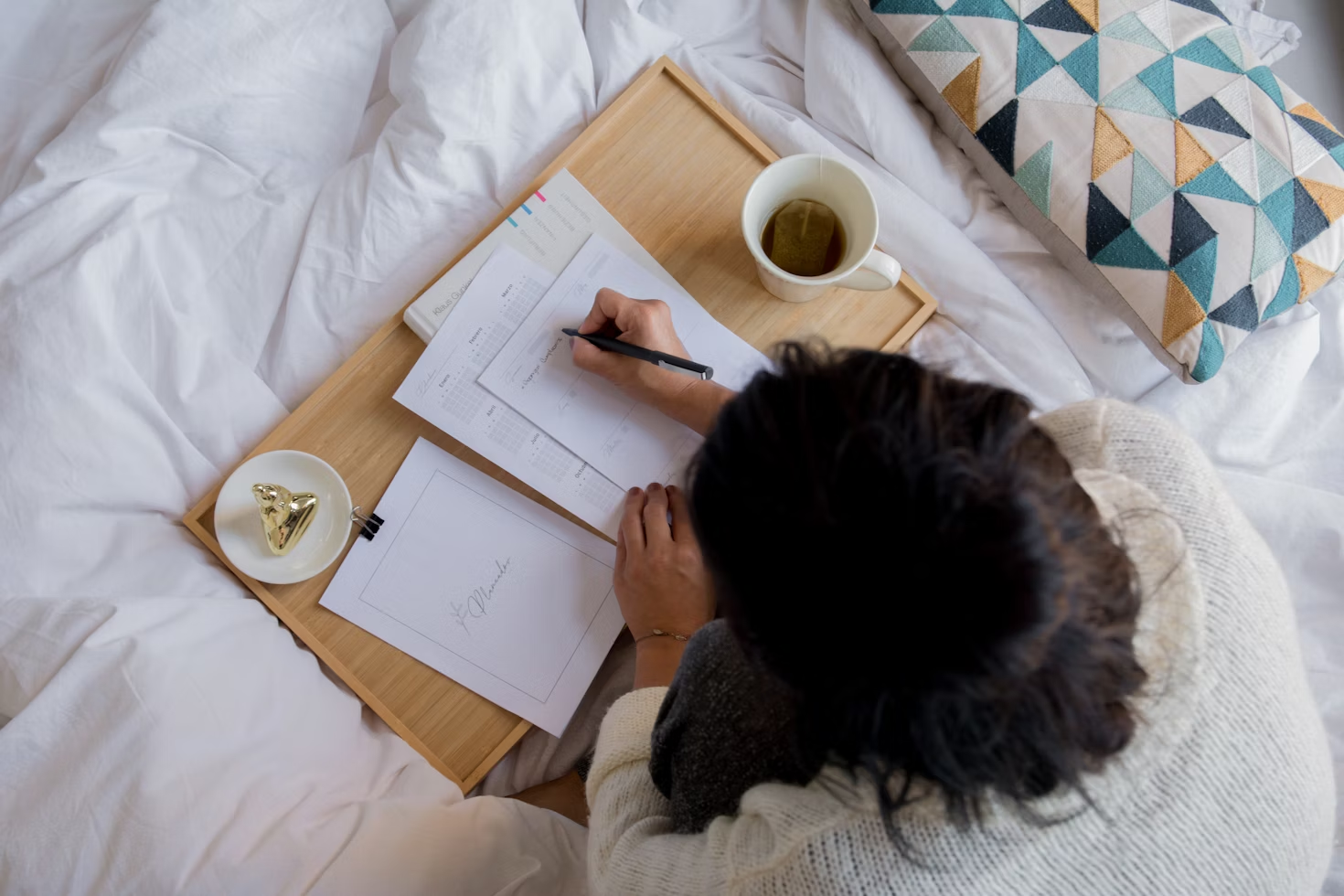 Femme écrivant sur du papier blanc sur un plateau en bois posé sur son lit avec une tasse de thé posée dessus