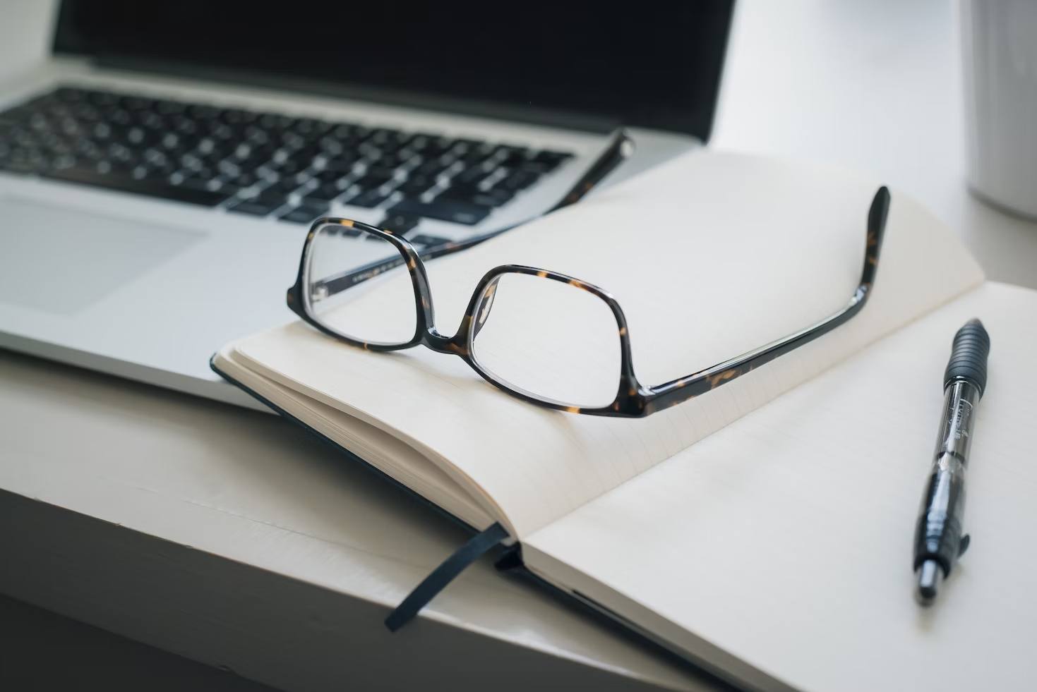 Lunettes de vue à montre foncée posées sur un carnet à côté d'un stylo bille le tout posé sur un ordinateur portable sur un bureau