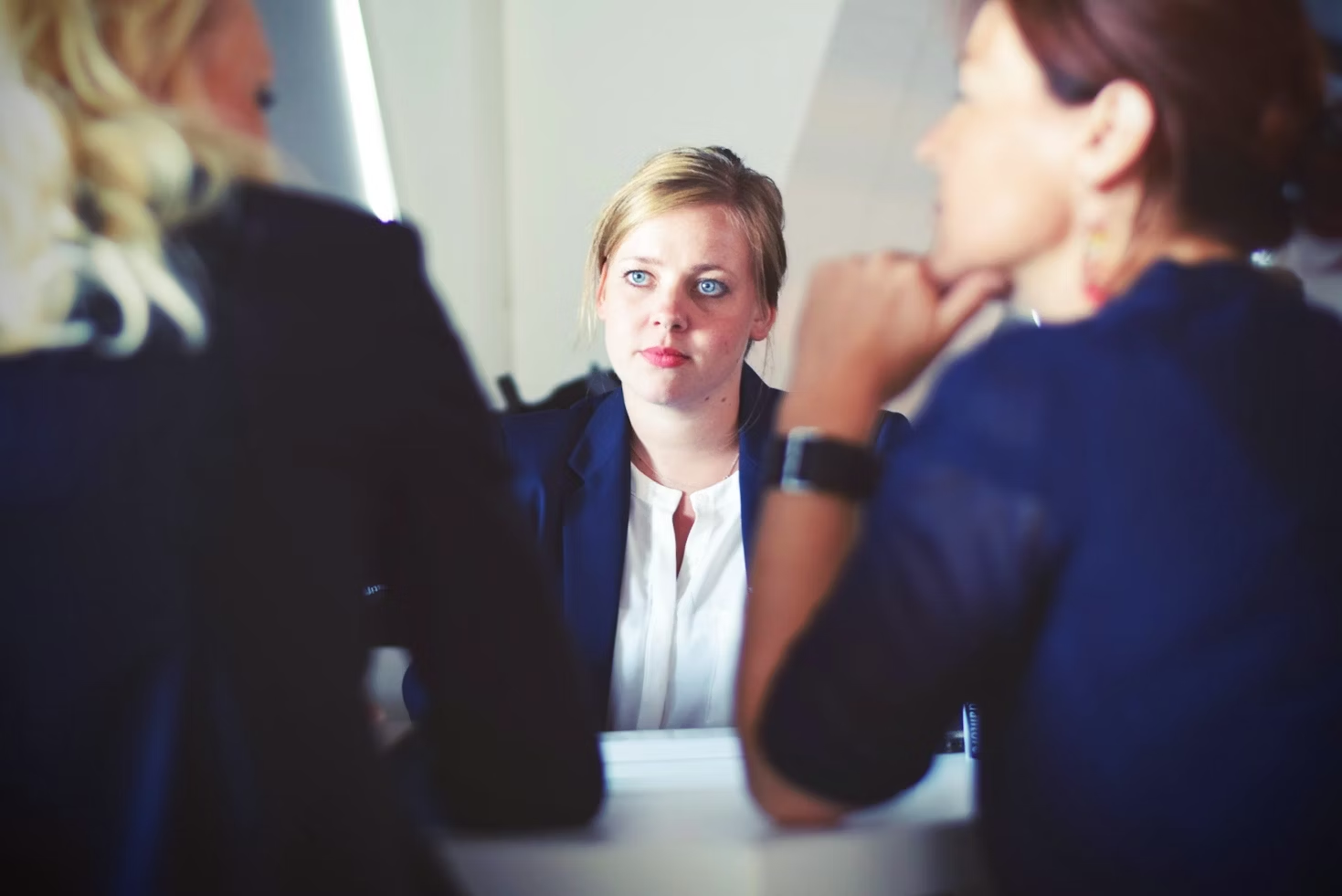 Femme blonde aux yeux bleus portant un blazer bleu marine faisant face à deux femmes de dos assises à une table