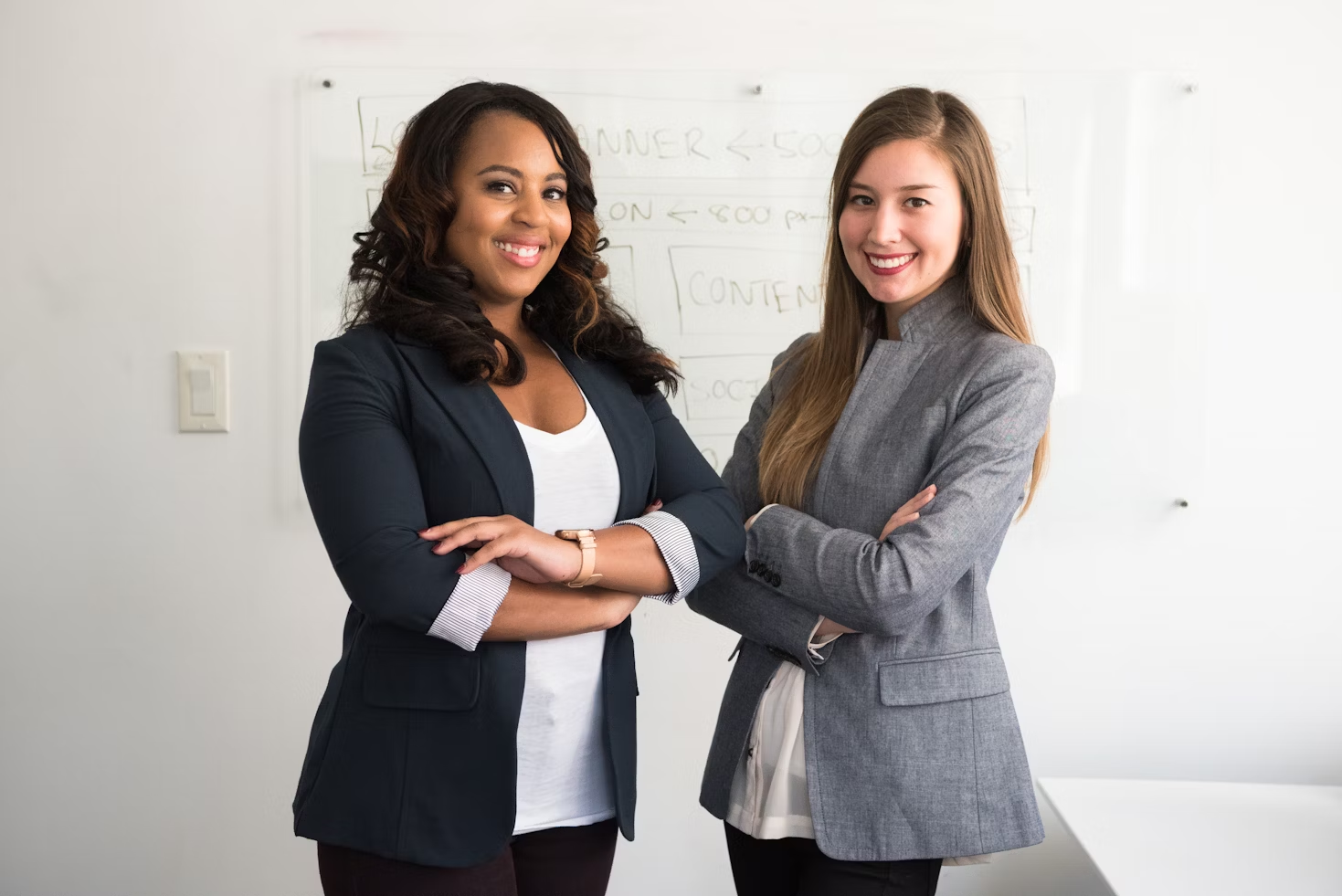 Deux femmes en blazer bleu foncé et gris debout les bras croisés devant un tableau blanc avec un schéma