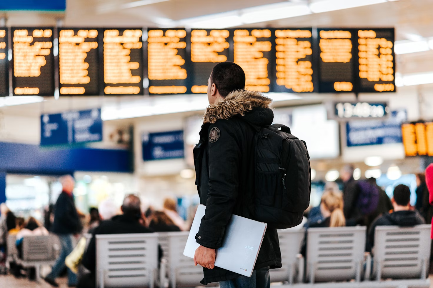 Homme debout dans un terminal d'aéroport en parka portant un sac à dos noir et un ordinateur sous son bras