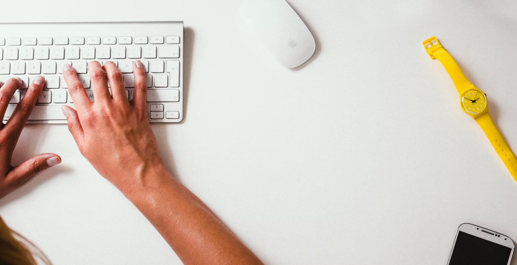 Mains pianotant sur un clavier Apple sans fil à côté d'une souris Apple sans fil avec un téléphone et une montre jaune posés sur un fond blanc