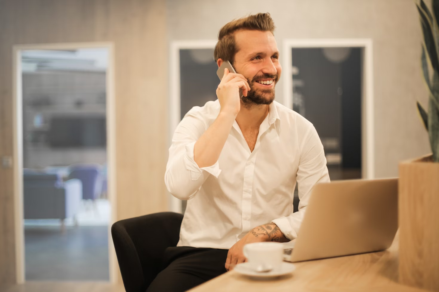 Homme en chemise blanche au téléphone assis à un bureau devant son ordinateur portable en souriant