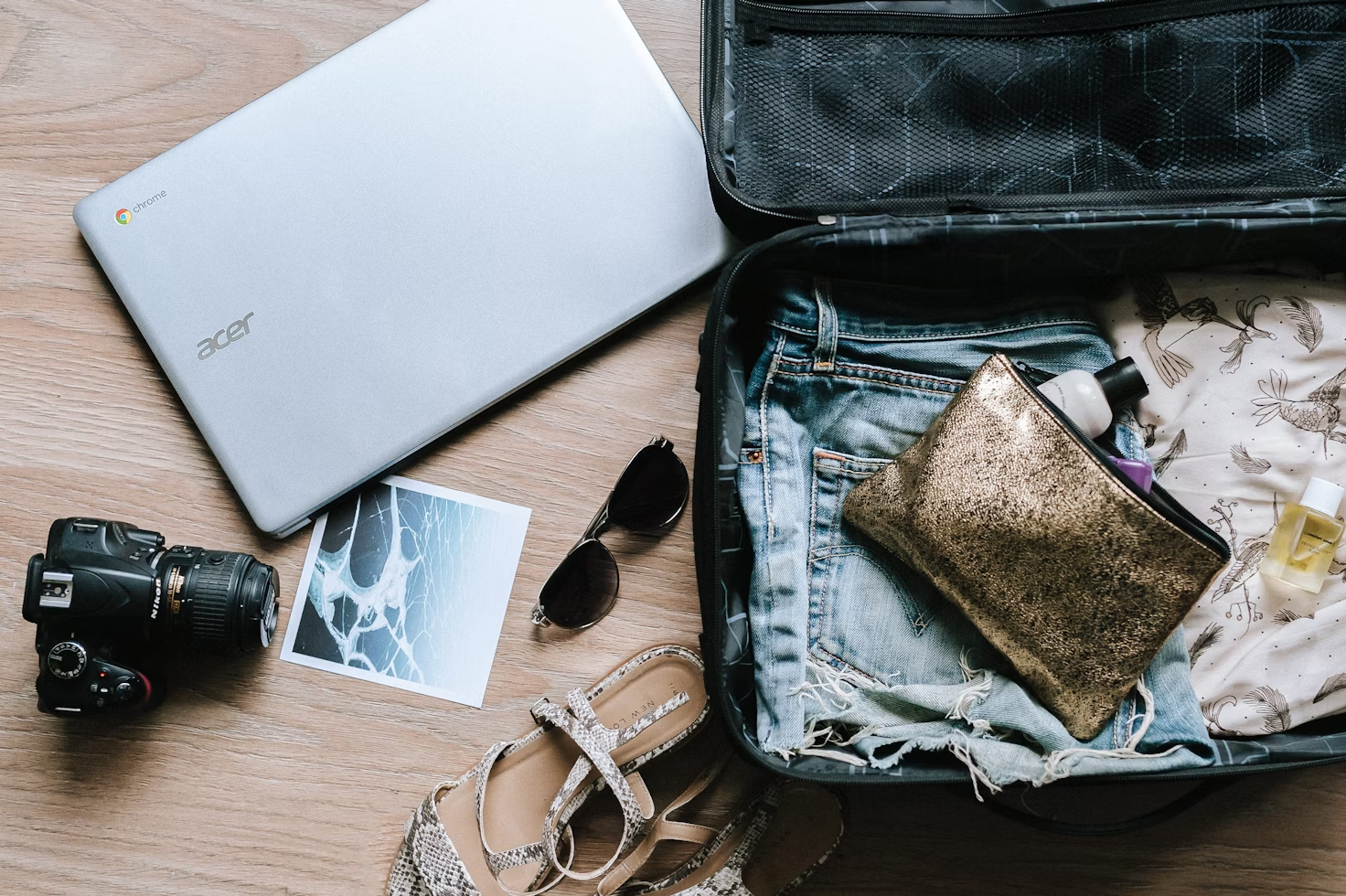 Valise posée sur le sol avec des habits et une trousse de toilette dorée posée à côté de chaussures d'un ordinateur et d'un appareil photo