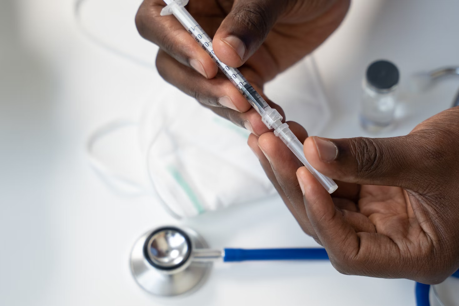 Personne tenant une seringue au dessus d'une table avec un stéthoscope bleu posé à côté d'un masque chirurgical